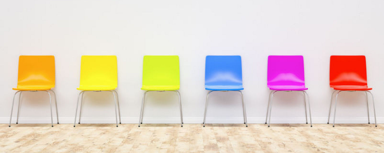 Six chairs, each one a different, bright color, in a row against a white wall.